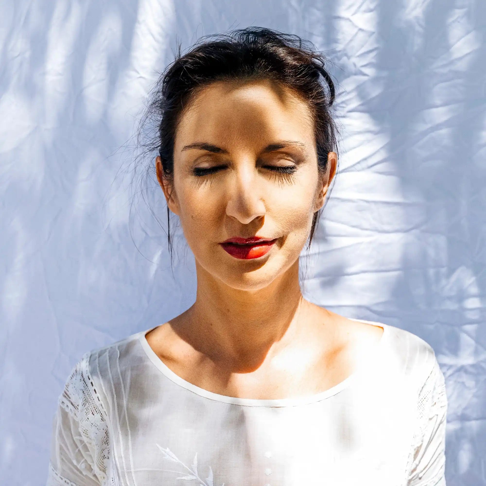 Portrait of someone wearing red lipstick and a white top with closed eyes in serene expression.